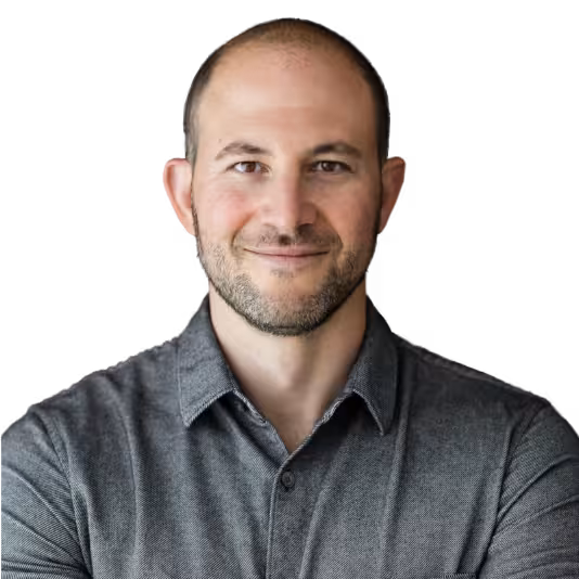 A man with short hair and a trimmed beard, wearing a grey collared shirt, stands with arms crossed and smiles at the camera against a plain white background.